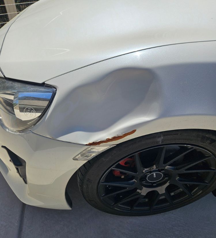 White sports car fender with large dent and rusted scrape above the wheel.